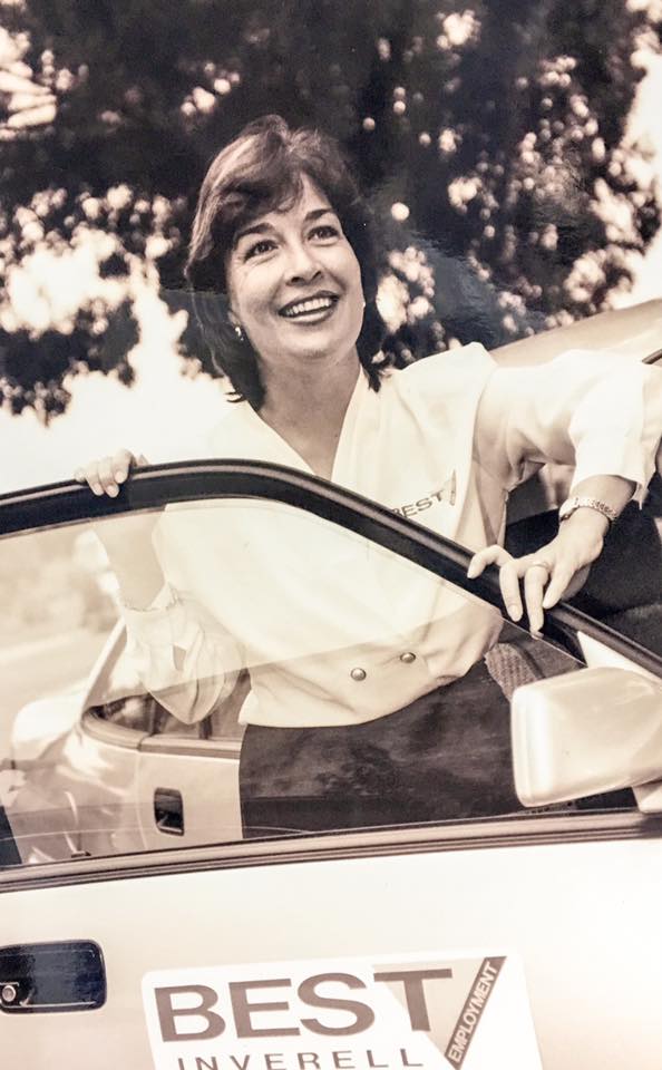 On old black and white image of Penny Alliston-Hall wearing a white shit with a BEST Employment logo, standing in the open door of an old car with the BEST Employment logo. 